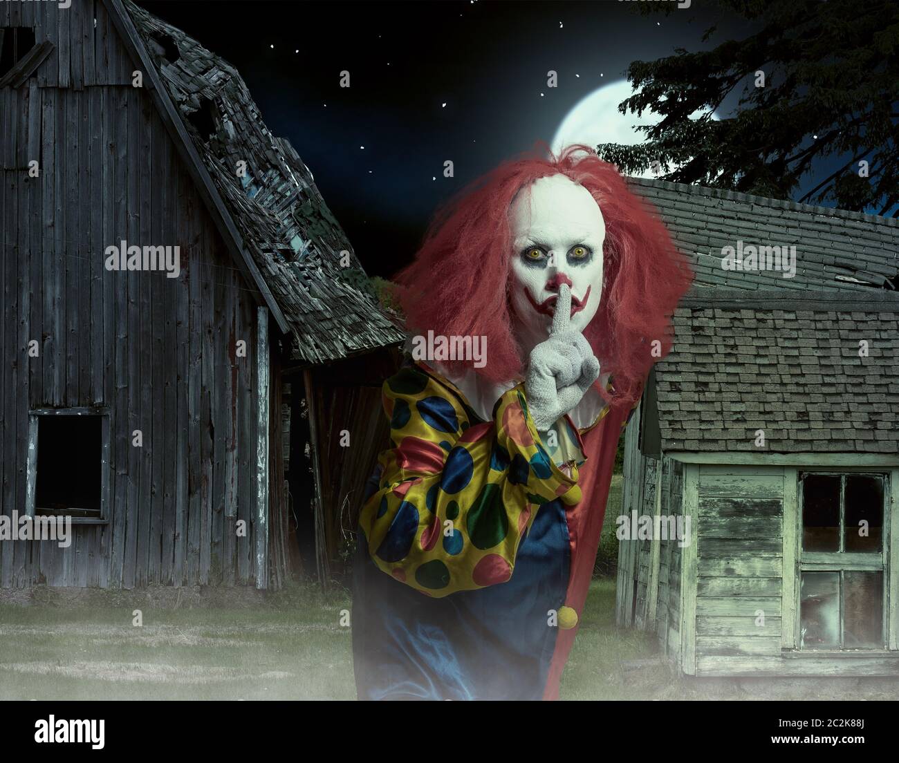 eerie clown with finger on mouth in front of a scary scene Stock Photo