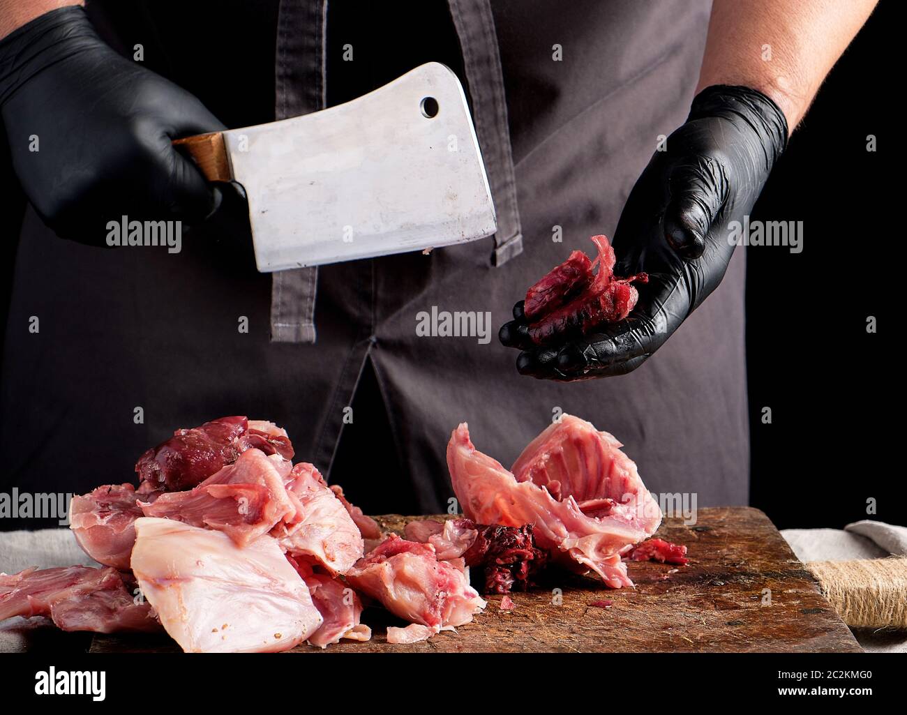 chef in black latex chunks cuts into pieces raw rabbit meat on a brown wooden board, cooking on a dark background Stock Photo