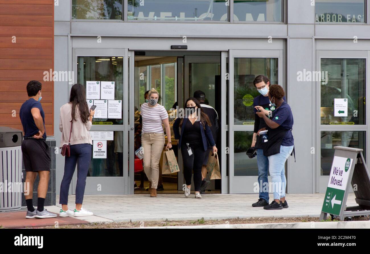 Baltimore, Maryland, USA. 17th June, 2020. Two female customers with face masks carrying paper bags of groceries emerge from the relocated Baltimore Harbor East Whole Foods Market location at 711 South Central Avenue on opening day while other patrons wait for permission from staff monitoring the doors to enter; a 'We've Moved' sign with logo beside them. The previous location of this branch of the Amazon-owned multinational supermarket chain, which closed a few days earlier, was located on nearby Fleet Street. Kay Howell/Alamy Live News Stock Photo