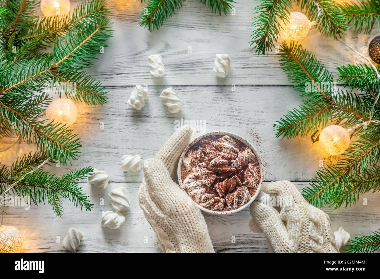 Woman's hands in wool gloves holding a cup of hot chocolate with marshmallows with decorated Christmas tree branches Stock Photo