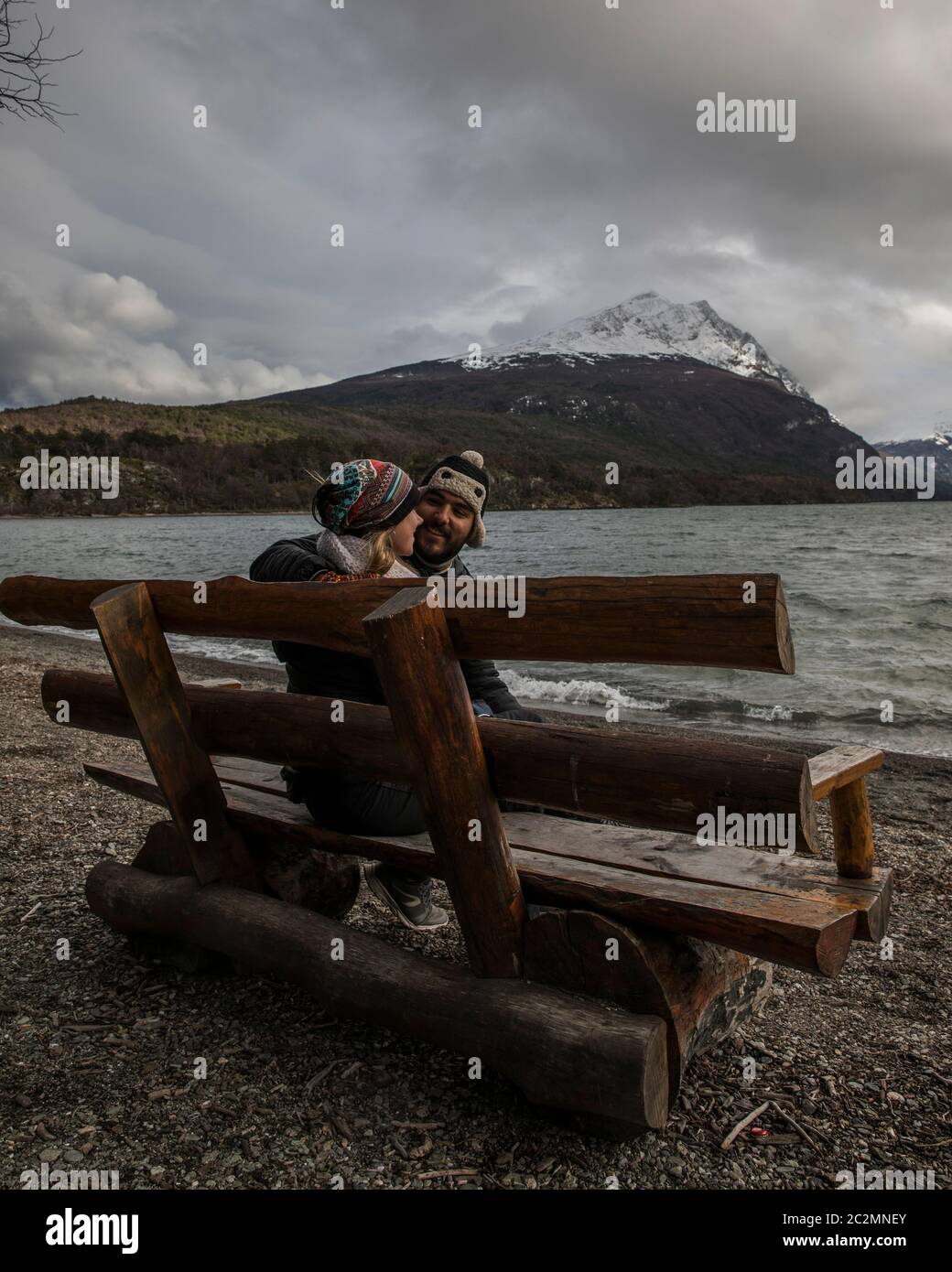 happy couple sitting in a bench in the beach in front of the ocean with a mountain with snow in the top in the background Stock Photo