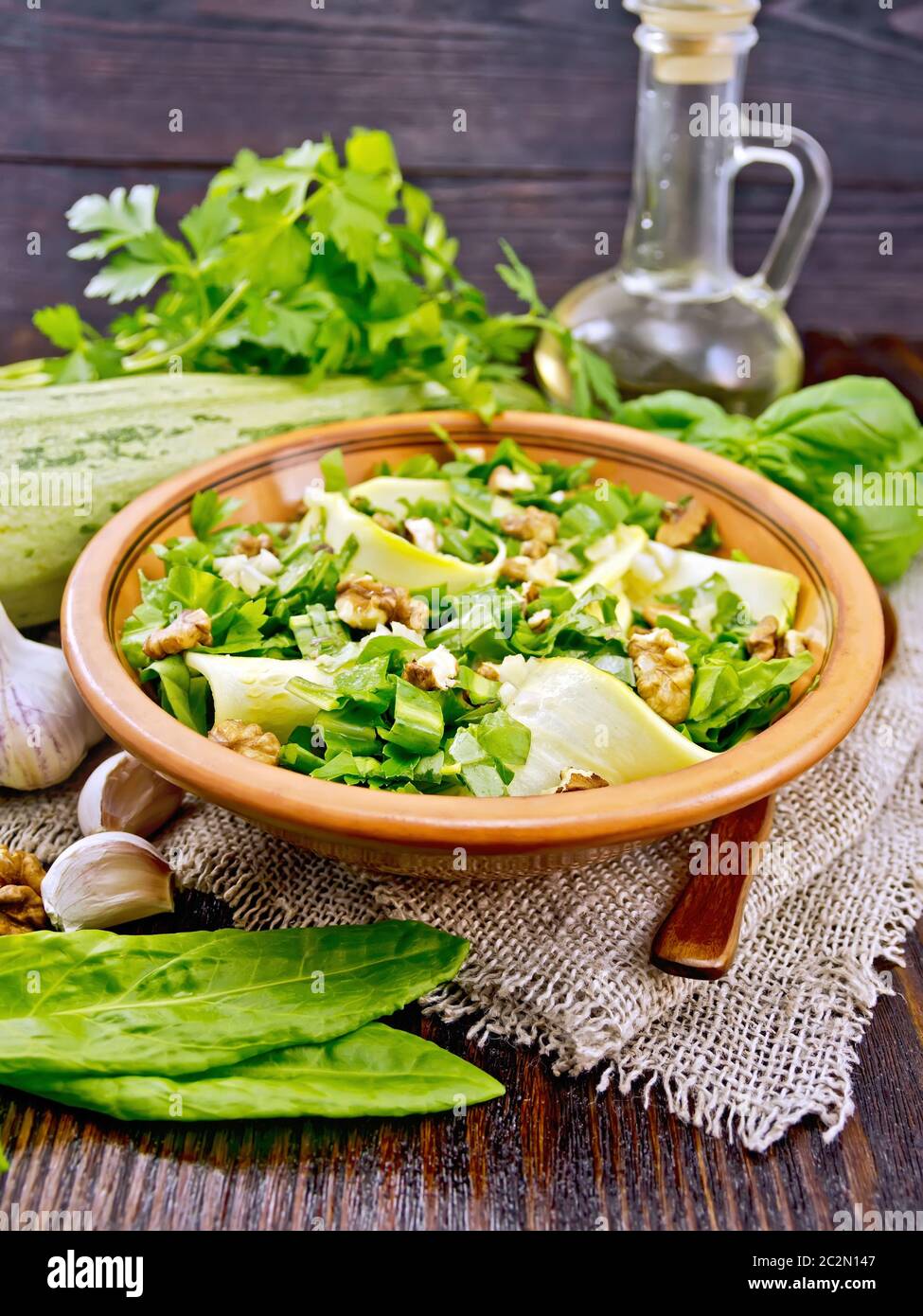 Salad of young zucchini, sorrel, garlic and nuts, seasoned with vegetable oil in clay plate on napkin of sackcloth on a dark wooden board background Stock Photo