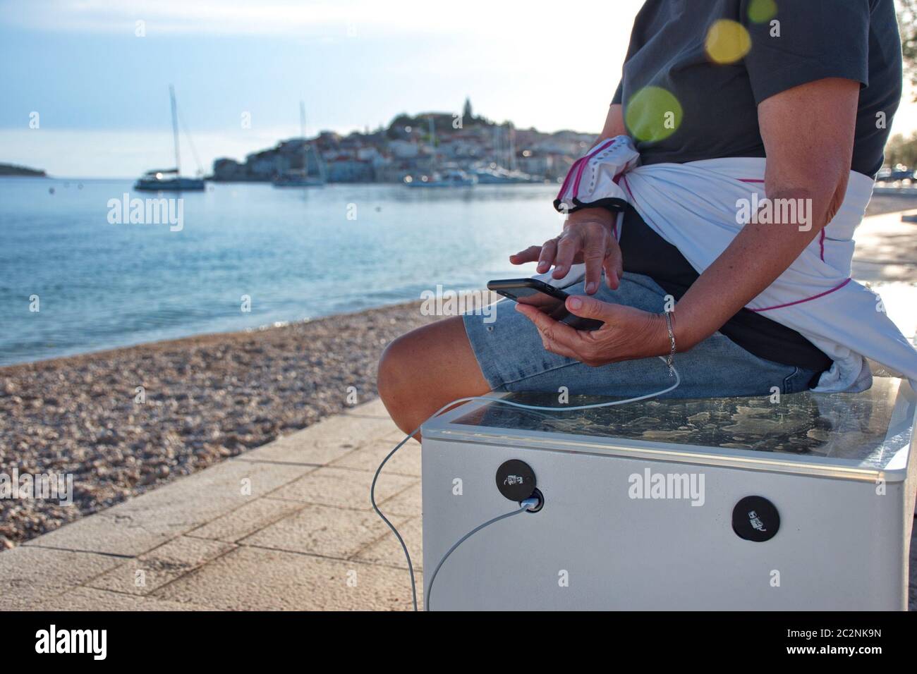Midsection of woman sitting on smart bench charging mobile phone with cityscape of Mediterranean town in background Stock Photo