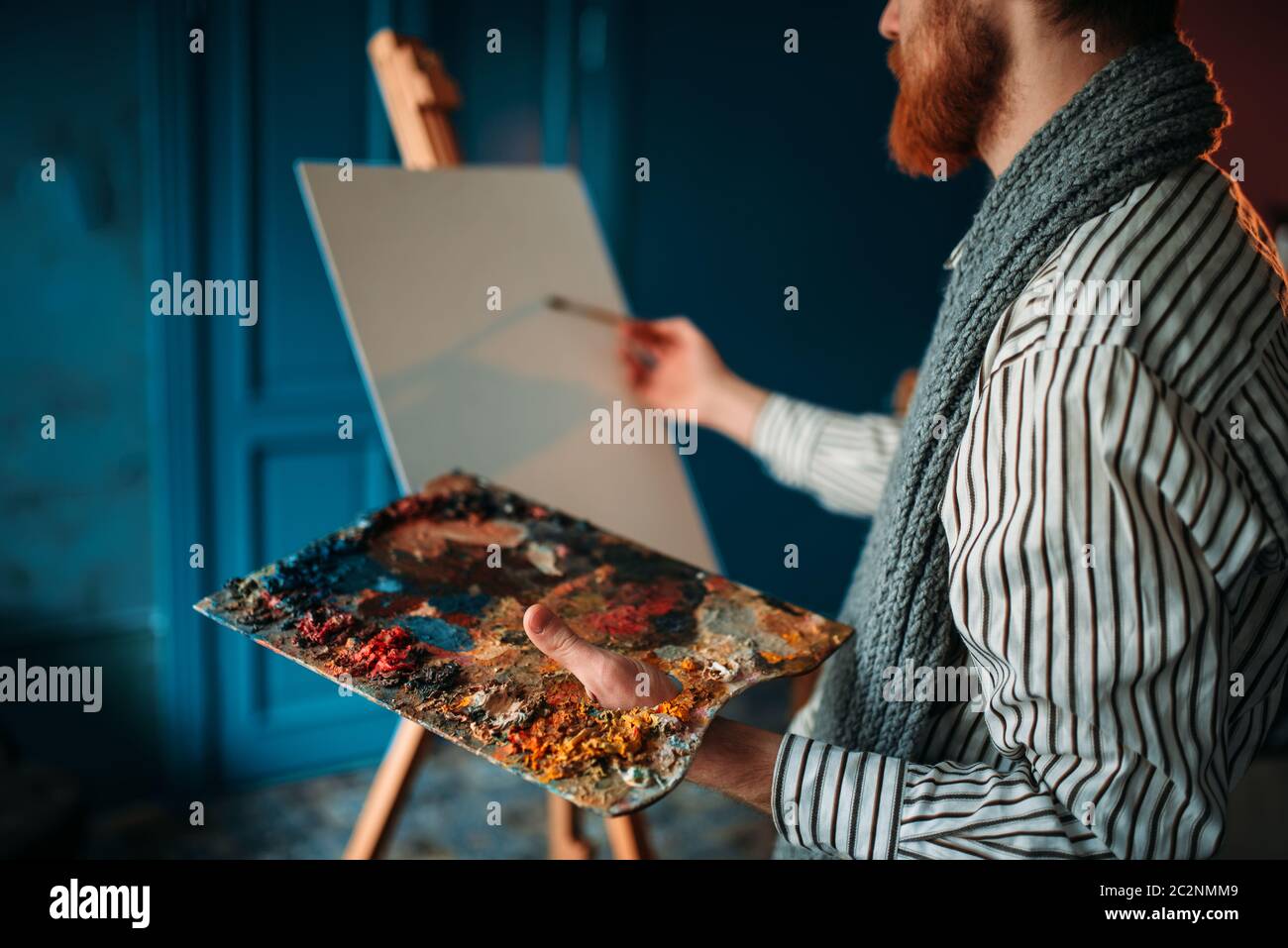 Male artist with palette and brush in hand in front of easel. Oil paint ...