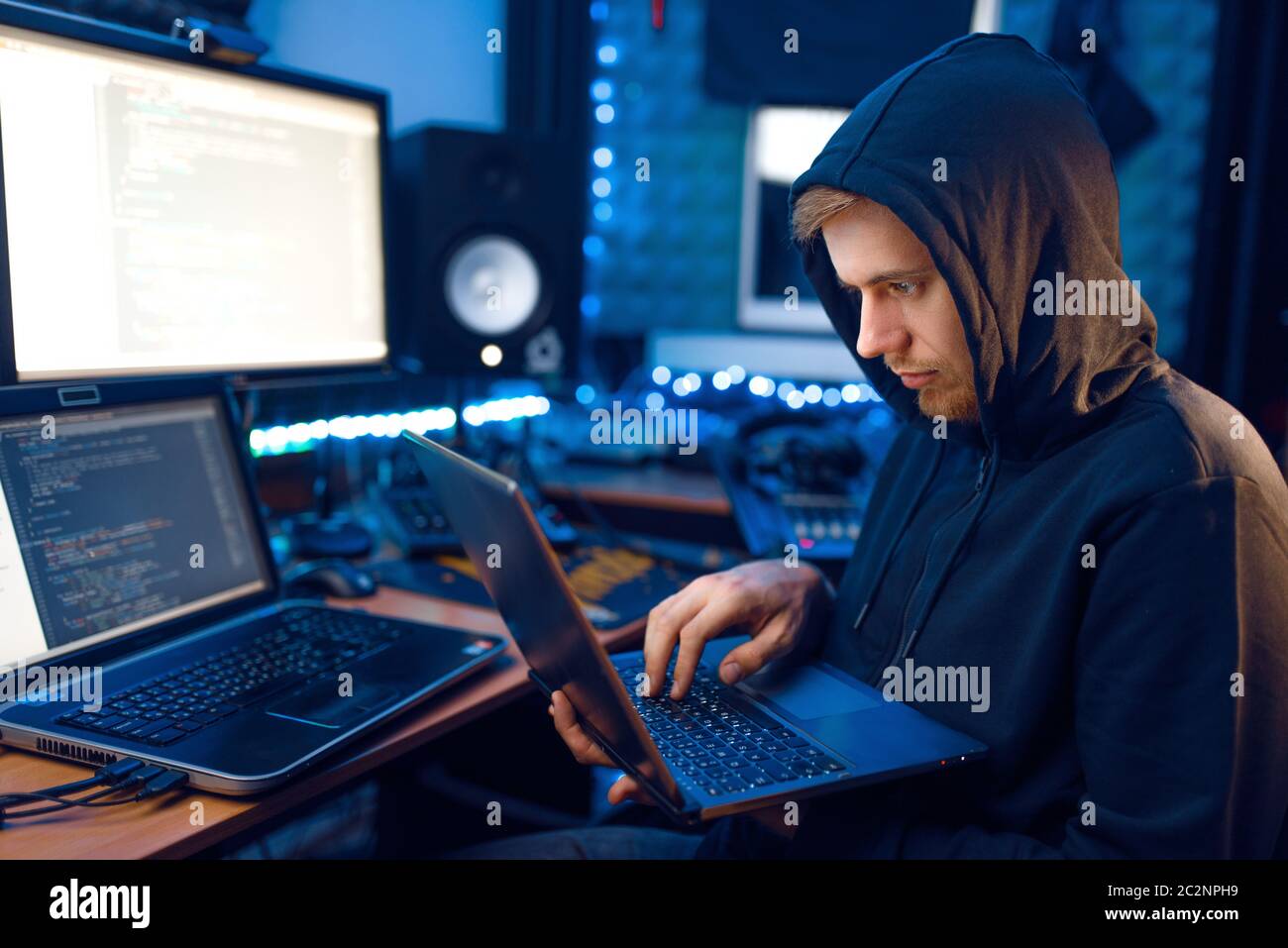 Hacker in hood shows thumbs up at his workplace with laptop and PC ...