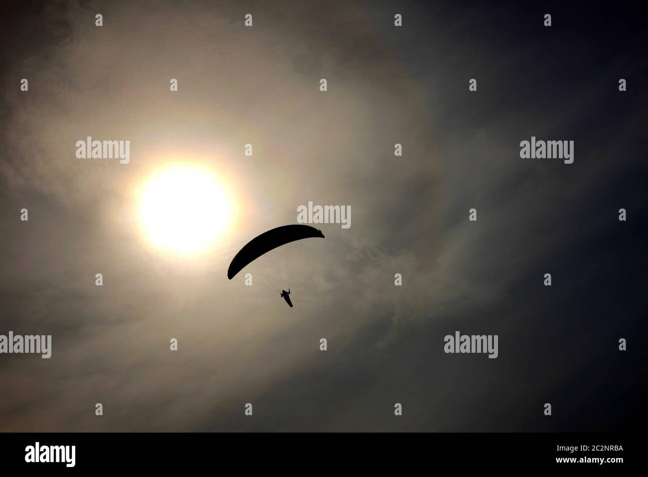 Paraglider soaring on wind thermals off Compton Isle of Wight in silhouette bright sun behind in grey cloudy sky Stock Photo
