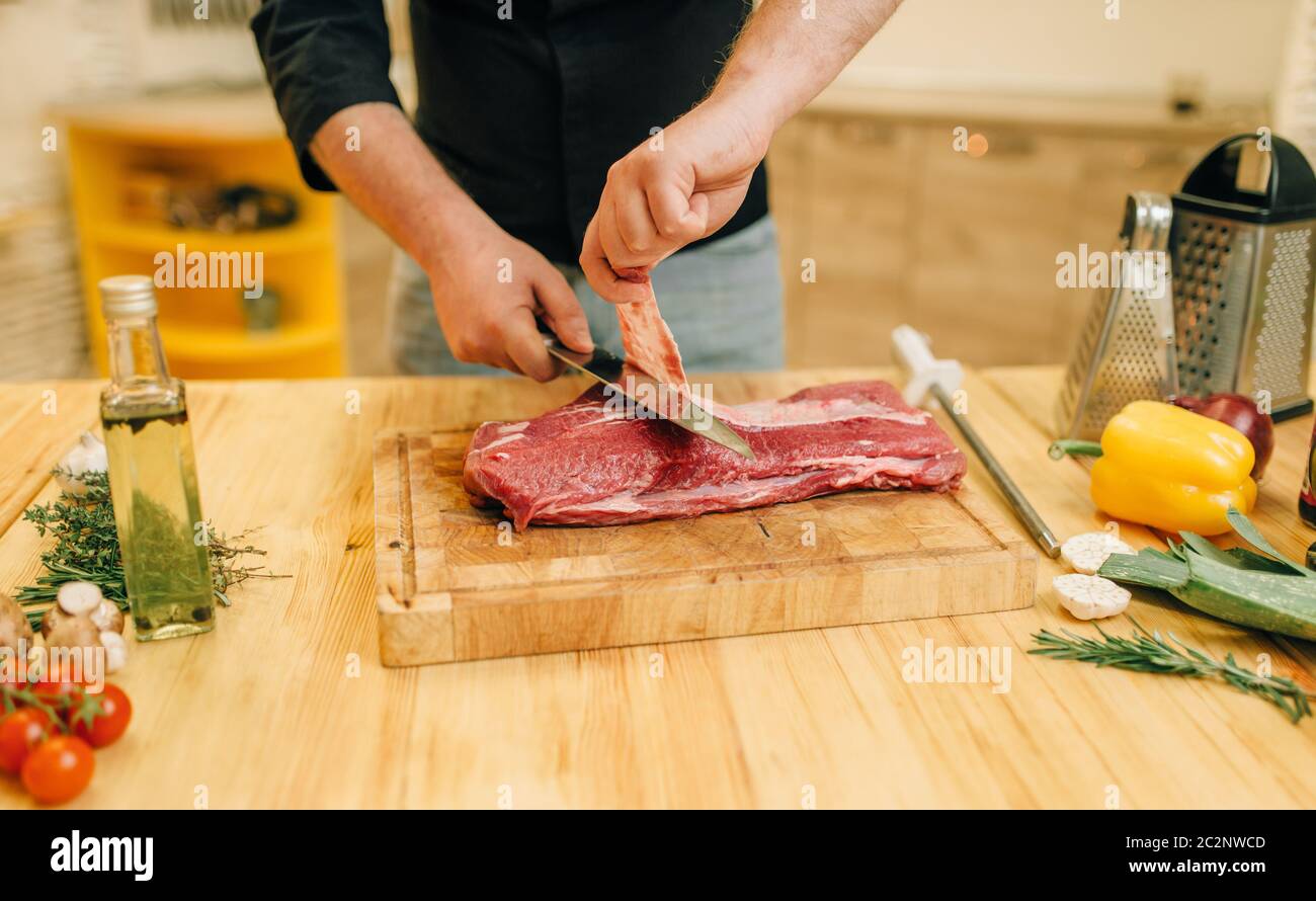 https://c8.alamy.com/comp/2C2NWCD/male-person-with-knife-cuts-raw-meat-on-wooden-board-kitchen-interior-on-background-chef-cooking-tenderloin-with-vegetables-spices-and-herbs-2C2NWCD.jpg