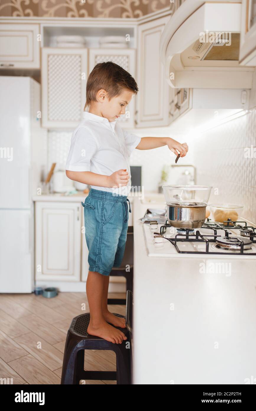 Litte boy cooking melt chocolate on the kitchen. Happy child standing on a chair at the stove and prepares sweet dessert Stock Photo