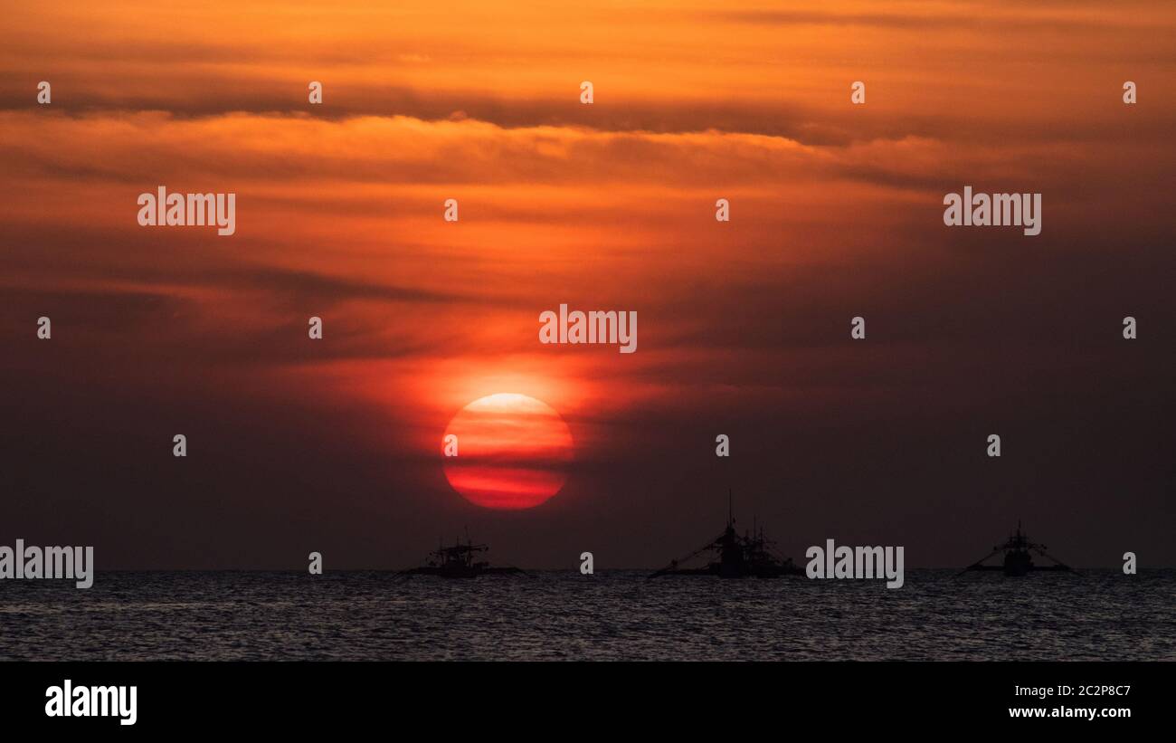 Thre boats lined up while the sun sets with its rays spreading upwards becoming dusk before darkness comes and it becomes night. Stock Photo