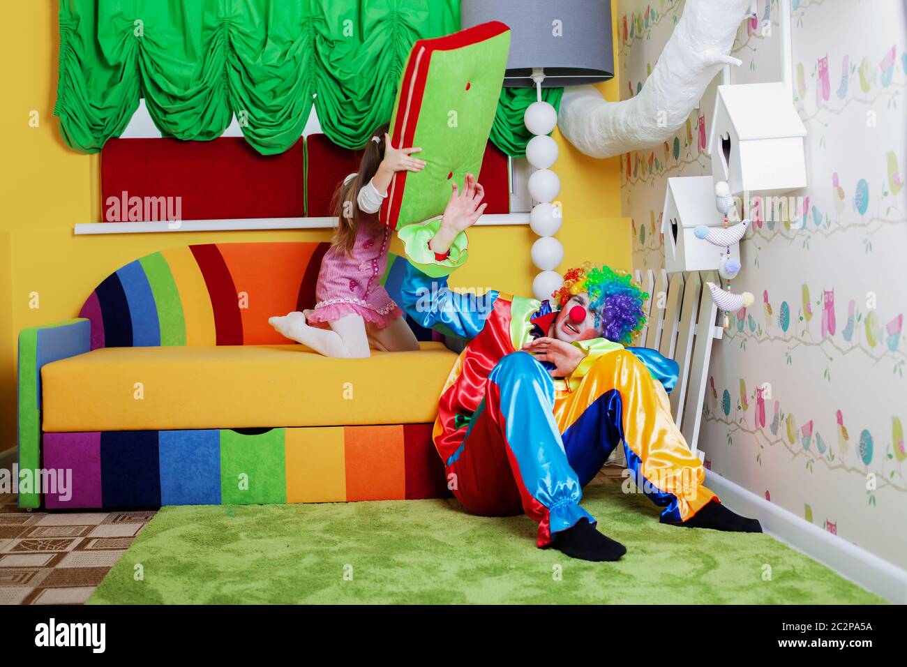 Happy little girl beats clown with a big pillow. with colourful sofa on the background Stock Alamy