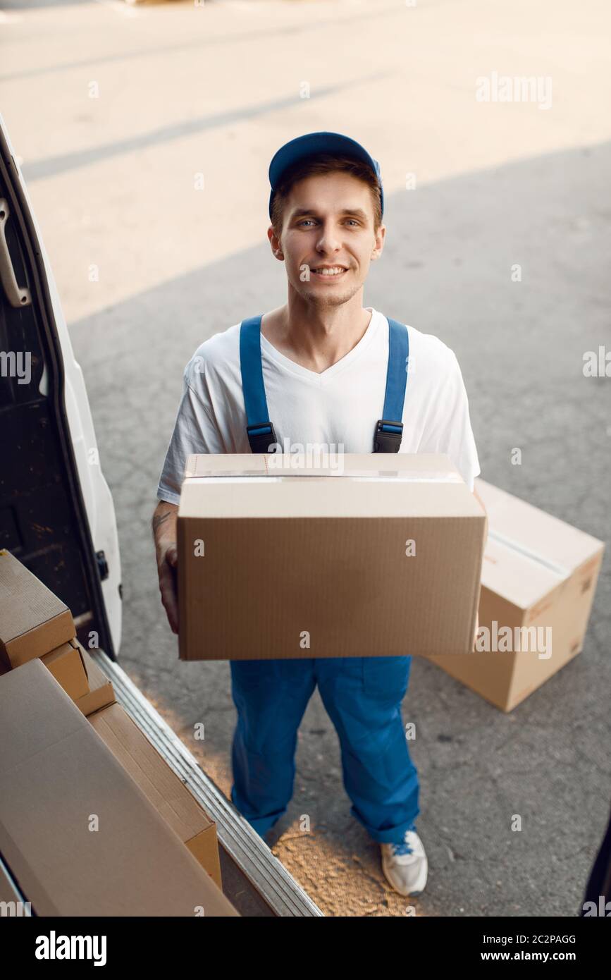 Deliveryman in uniform unloads the car with parcels, delivery service. Man standing at cardboard packages in vehicle, male deliver, courier or shippin Stock Photo