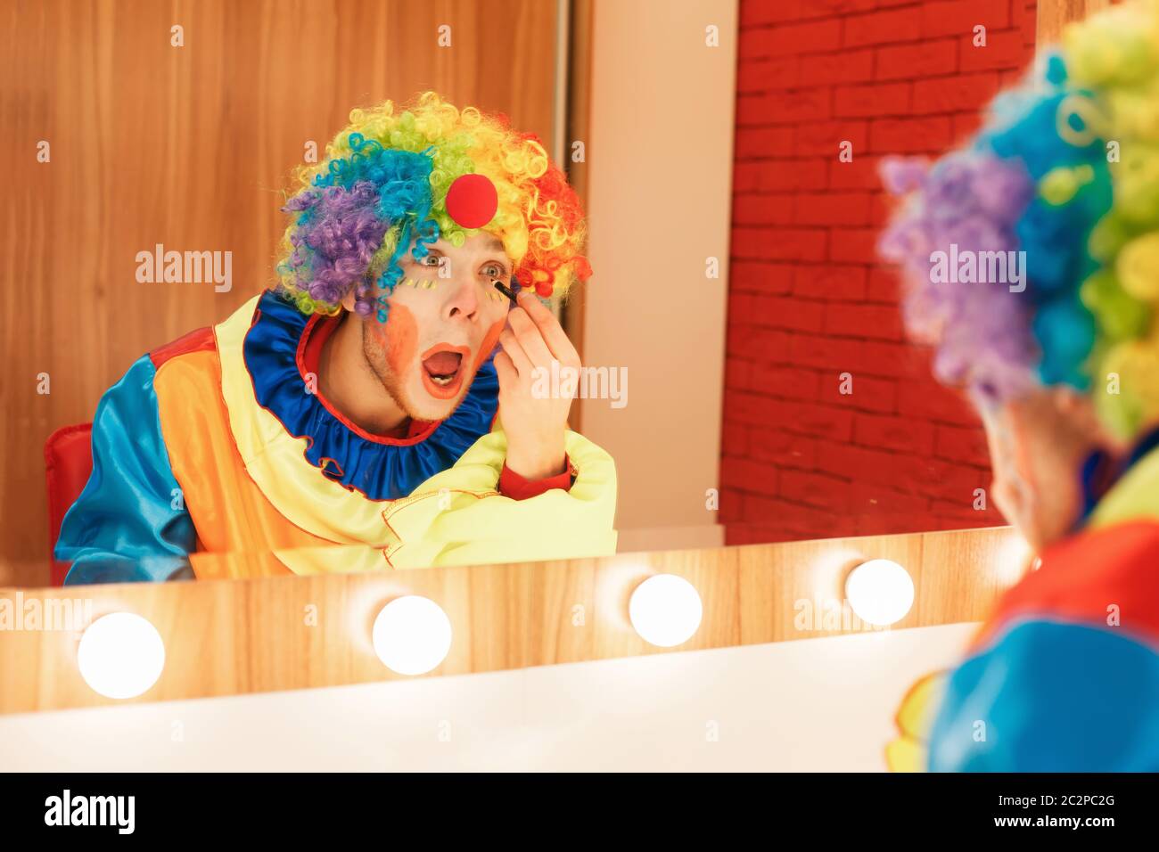Cheerful clown does a make-up in front of the mirror. Makeup room at ...