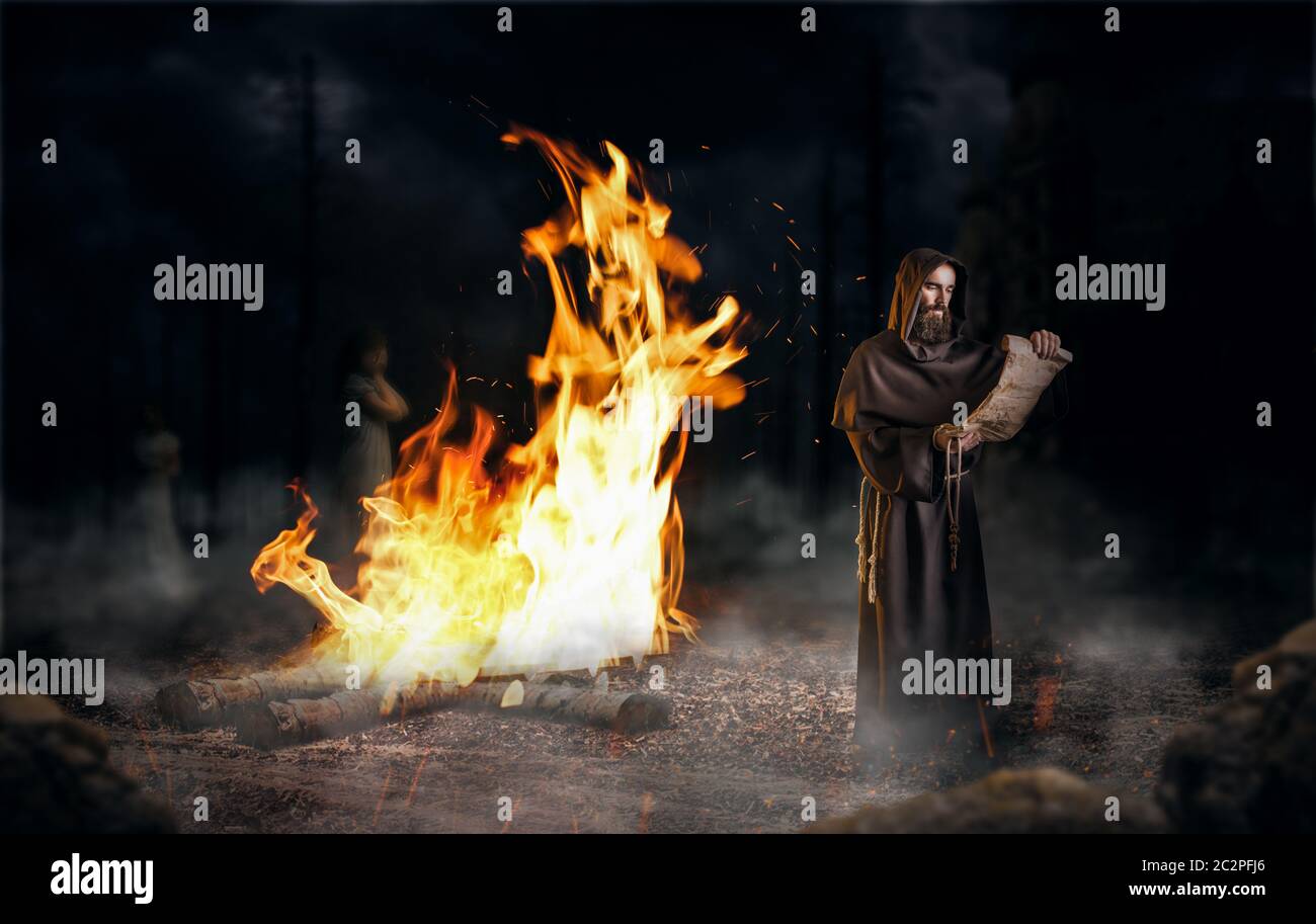 Medieval monk reads a prayer in the ancient manuscript against a big fire, religion. Mysterious friar in dark cape. Mystery and spirituality Stock Photo