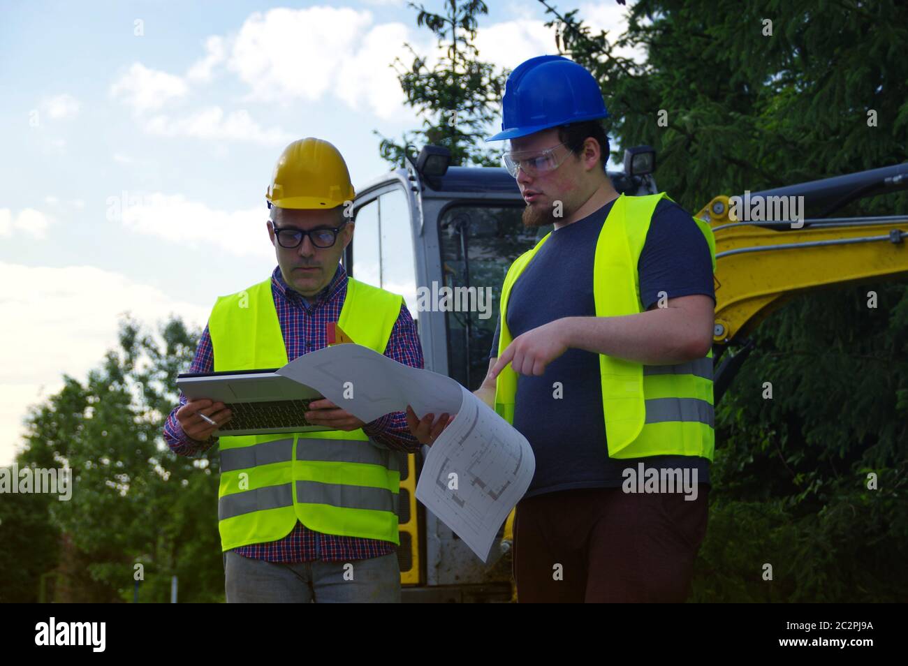 Engineer and worker on construction site with excavator in the background. A professional team working at the streets building. Two men in helmets tal Stock Photo