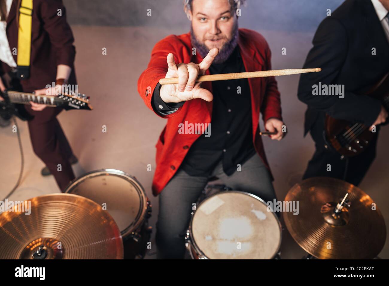 Drummer in red suit, performing on the stage of night club, electro and bas-guitarists on background. Rock band concert, music show Stock Photo