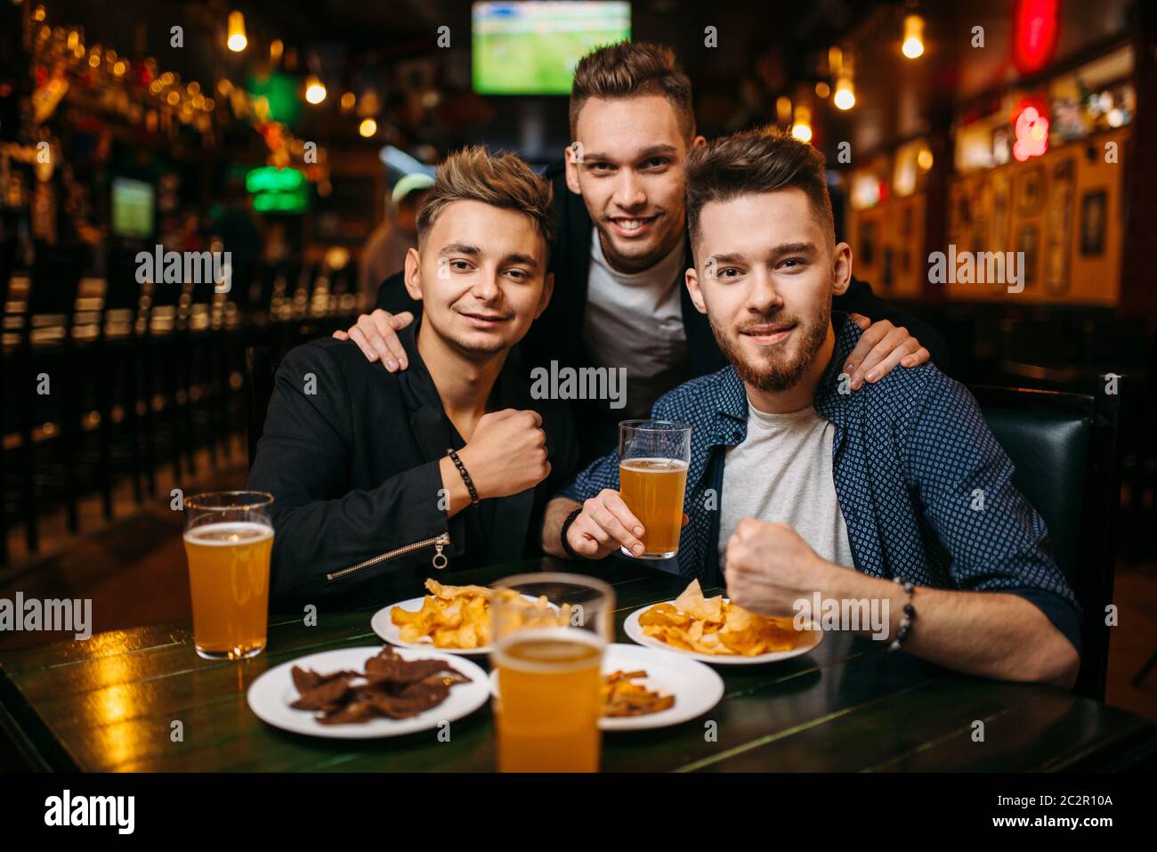 Three young men poses at the table with beer, crisps and crackers