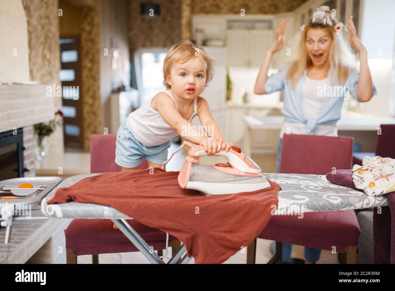 Little baby irons clothes on the kitchen, housewife in horror. Kid