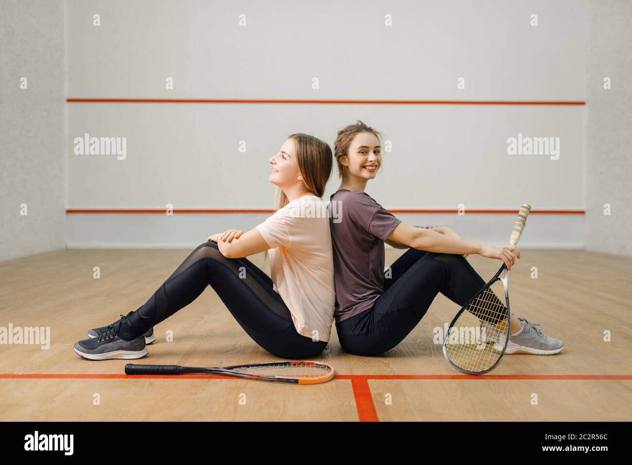 Two female players with squash rackets sits back to back on court floor