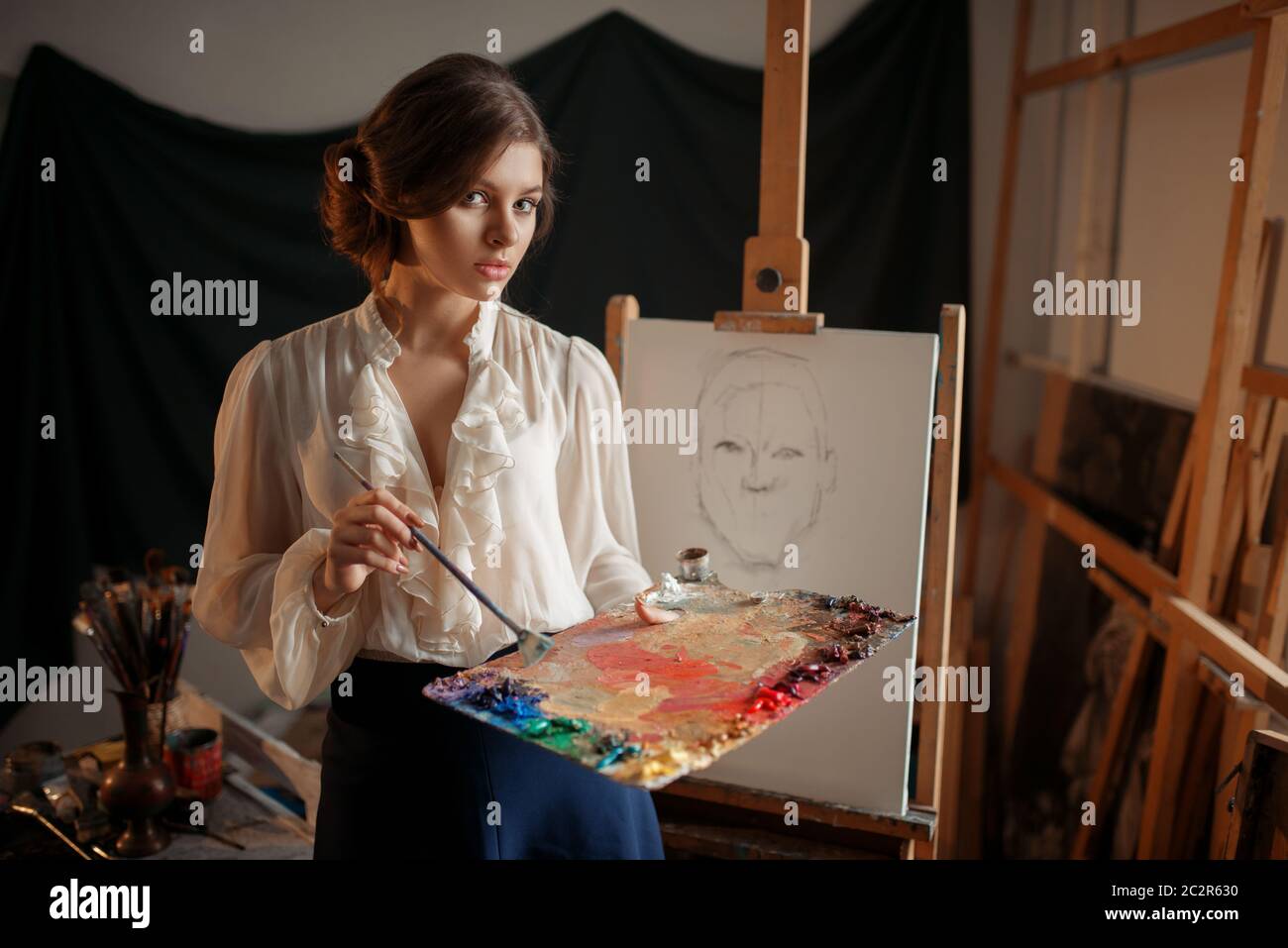 Female painter with color palette and brush standing against easel in ...