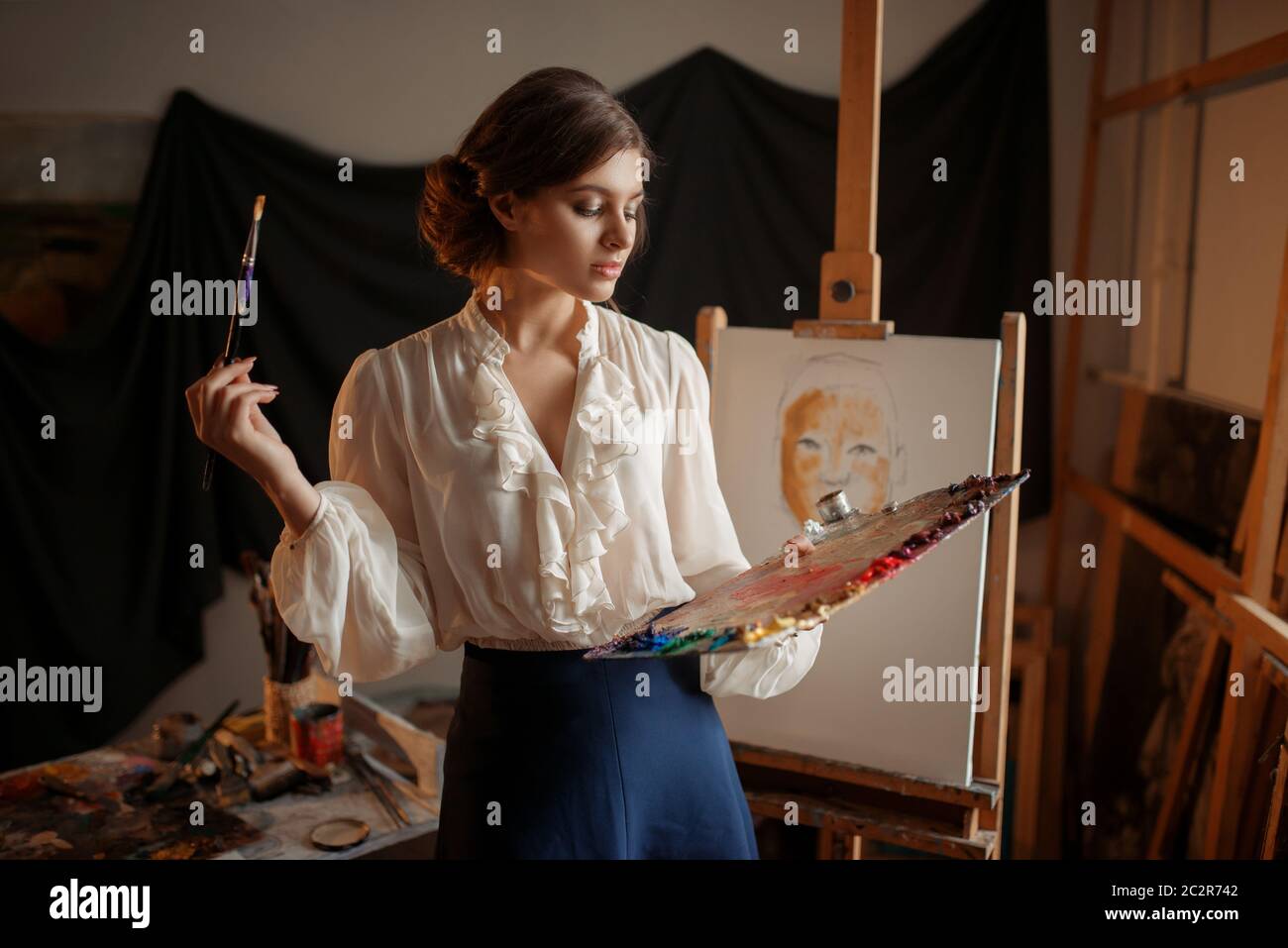 Female artist with color palette and brush standing against easel in ...