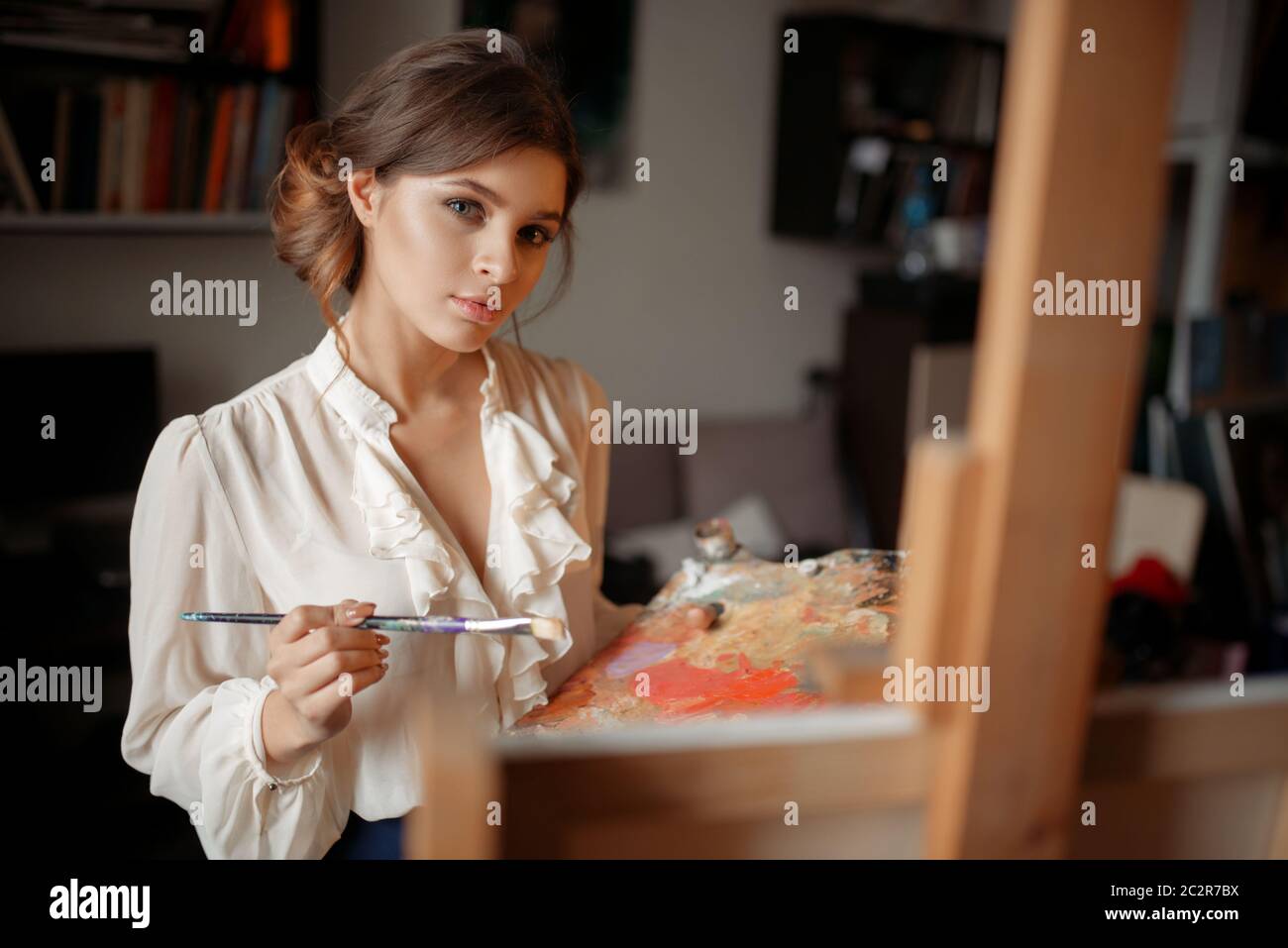 Thoughtful female painter with color palette and brush standing against ...