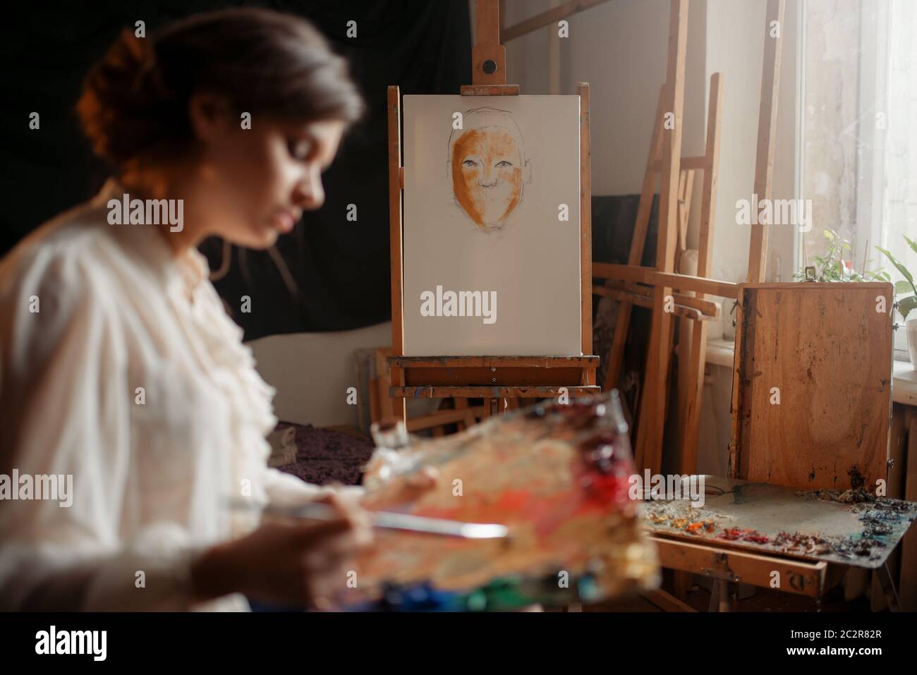 Female artist with color palette and brush standing against easel in ...