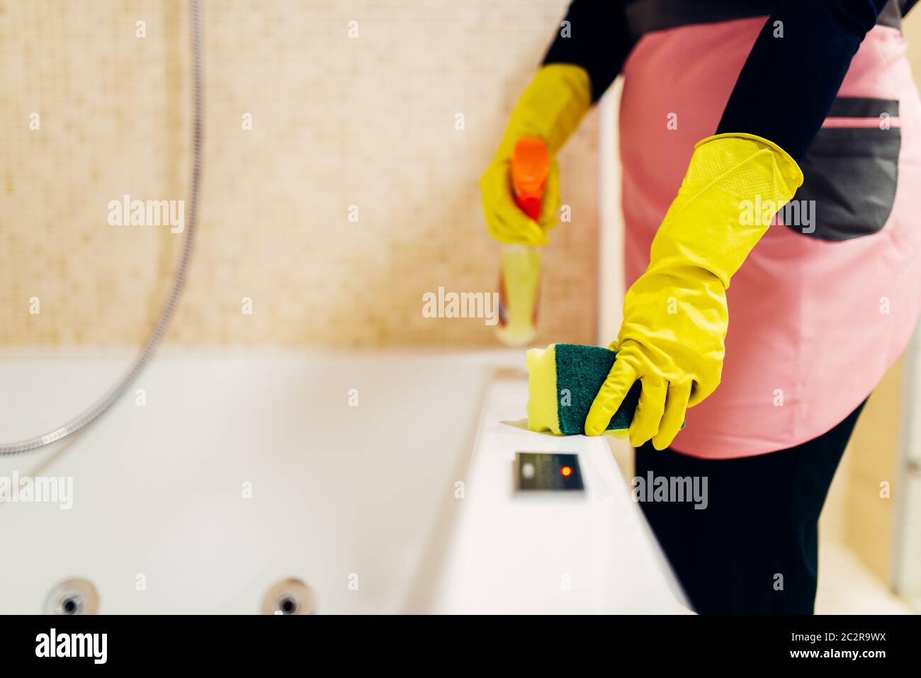 Maid hands in rubber gloves cleans the bathtube with a cleaning spray, hotel bathroom interior on background. Professional housekeeping service, charw Stock Photo