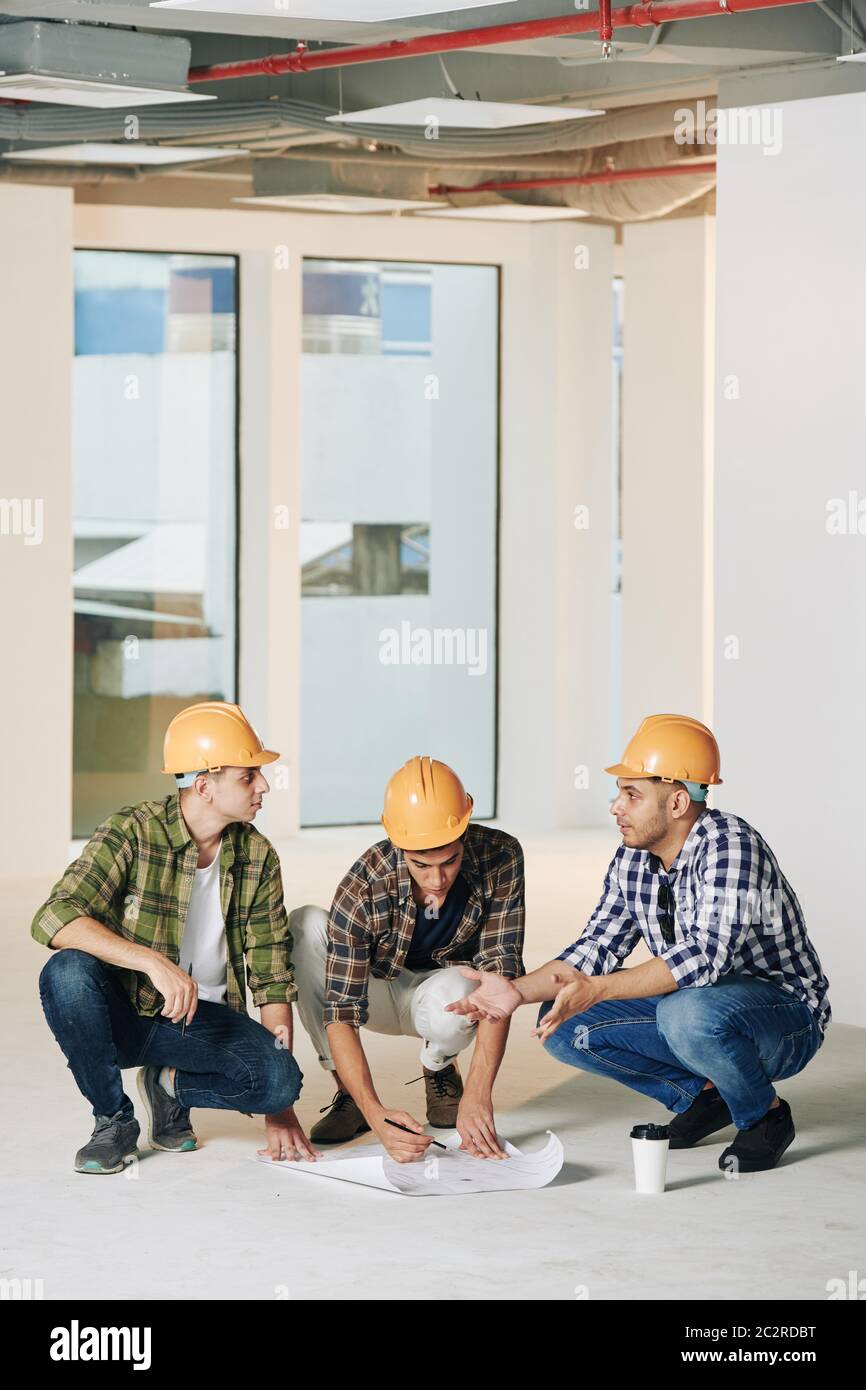 Vertical shot of three professional engineers sitting on hunkers in empty room of new building correcting construction plan Stock Photo