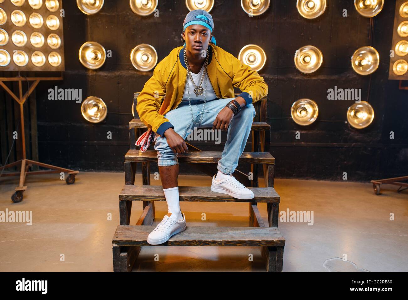 Black rapper in cap sitting on the steps, singer on stage with spotlights on background. Rap performer on scene with lights, underground music, urban Stock Photo