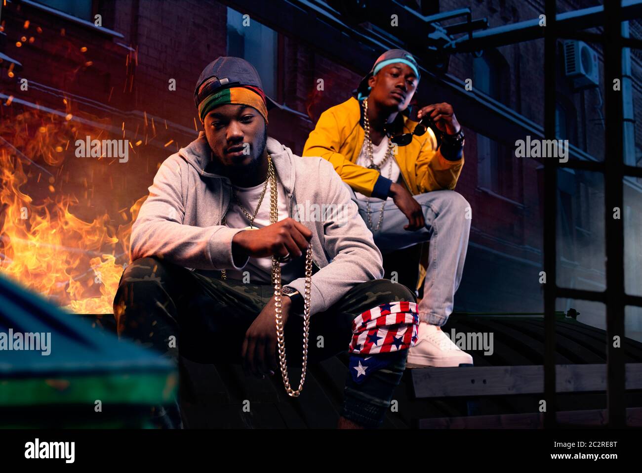 Two black rappers in caps sitting by the fire, night city street on background. Rap performers against cityscape, underground music concert, urban sty Stock Photo