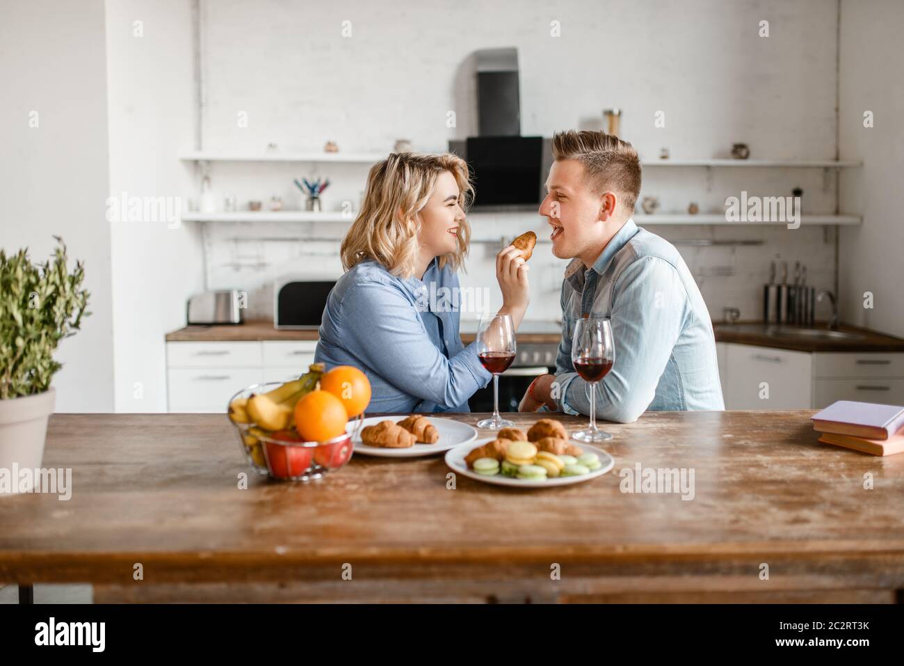 Love couple sitting at the table, romantic dinner. Man and woman drinks wine and eats fruits on the kitchen. Happy lifestyle, beautiful relationship Stock Photo