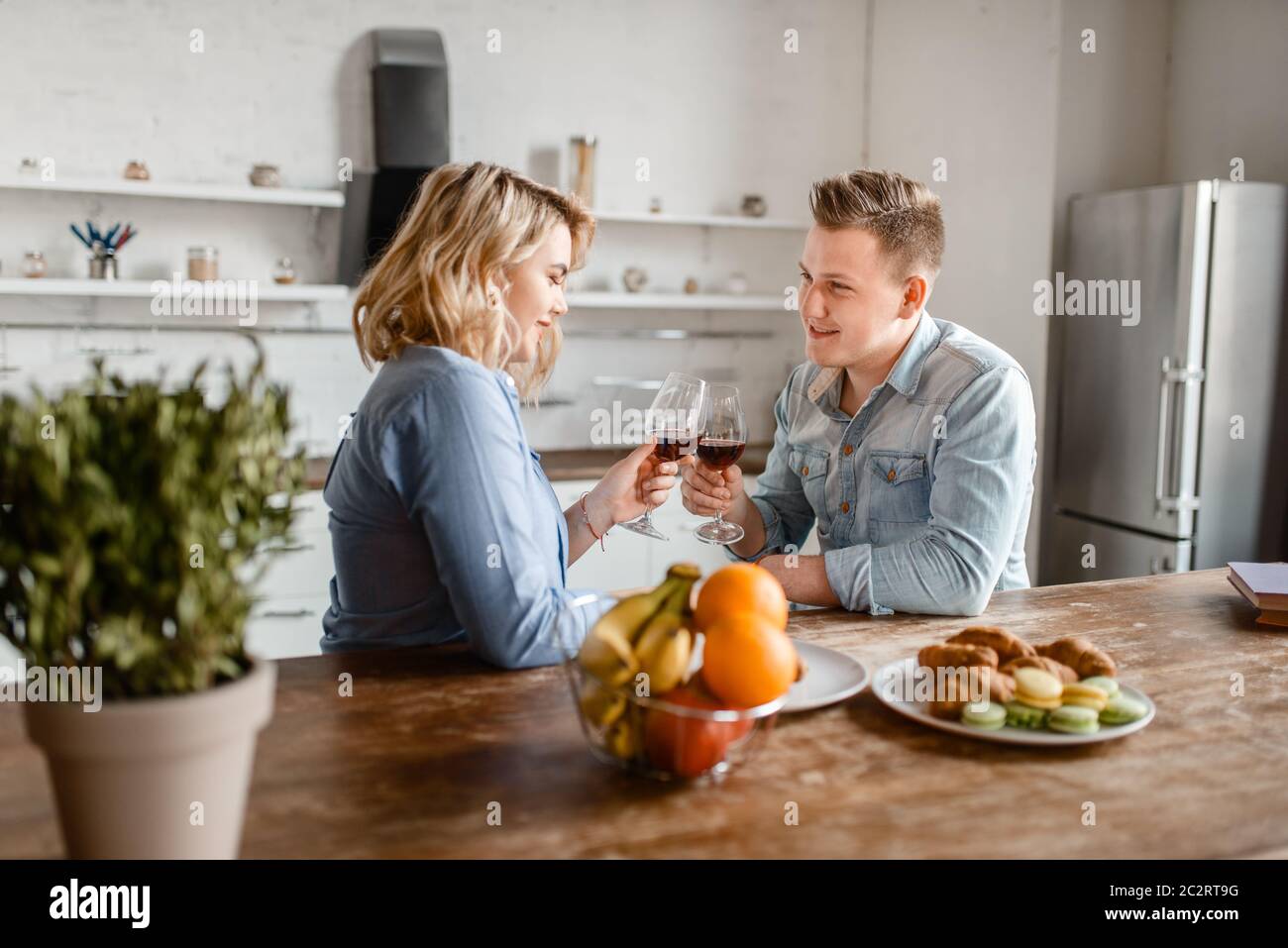 Love couple sitting at the table, romantic dinner. Man and woman drinks wine and eats fruits on the kitchen. Happy lifestyle, beautiful relationship Stock Photo