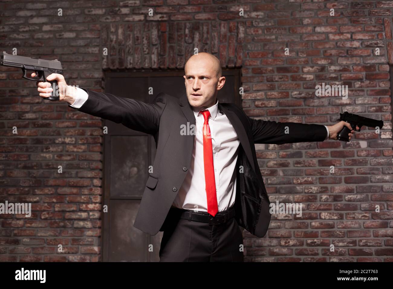 Serious contract killer shooting action wallpaper, background or poster. Bald assassin in red tie fires a pistols with two hands on city street. Profe Stock Photo