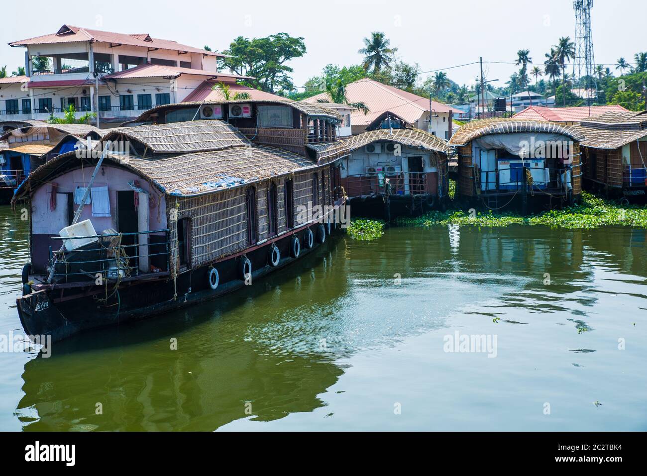 Small houses in a local village located next to Kerala's backwater on a bright sunny day and traditional Houseboat seen sailing through the river Stock Photo