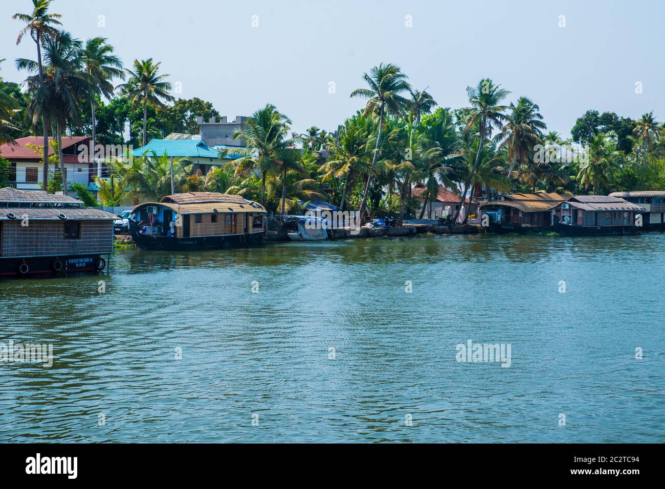 Small houses in a local village located next to Kerala's backwater on a bright sunny day and traditional Houseboat seen sailing through the river Stock Photo