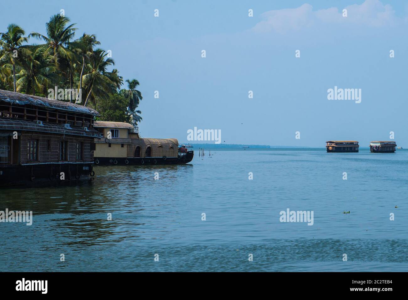 Small houses in a local village located next to Kerala's backwater on a bright sunny day and traditional Houseboat seen sailing through the river Stock Photo