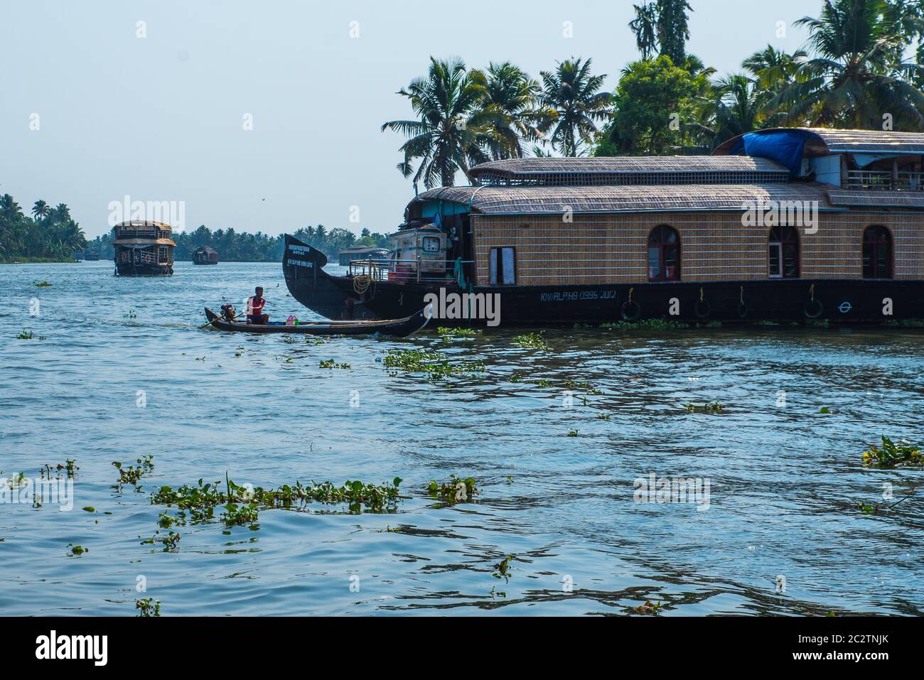 Small houses in a local village located next to Kerala's backwater on a bright sunny day and traditional Houseboat seen sailing through the river Stock Photo