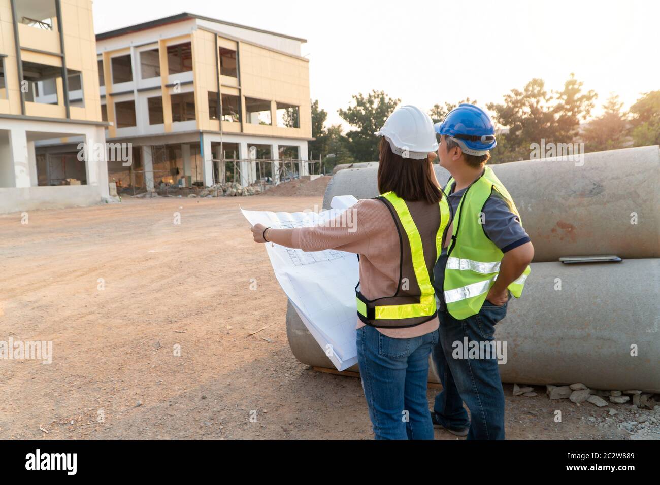 Asian woman construction engineer holding blueprint for checking construction site with technical engineer Building Location on the background, Concep Stock Photo
