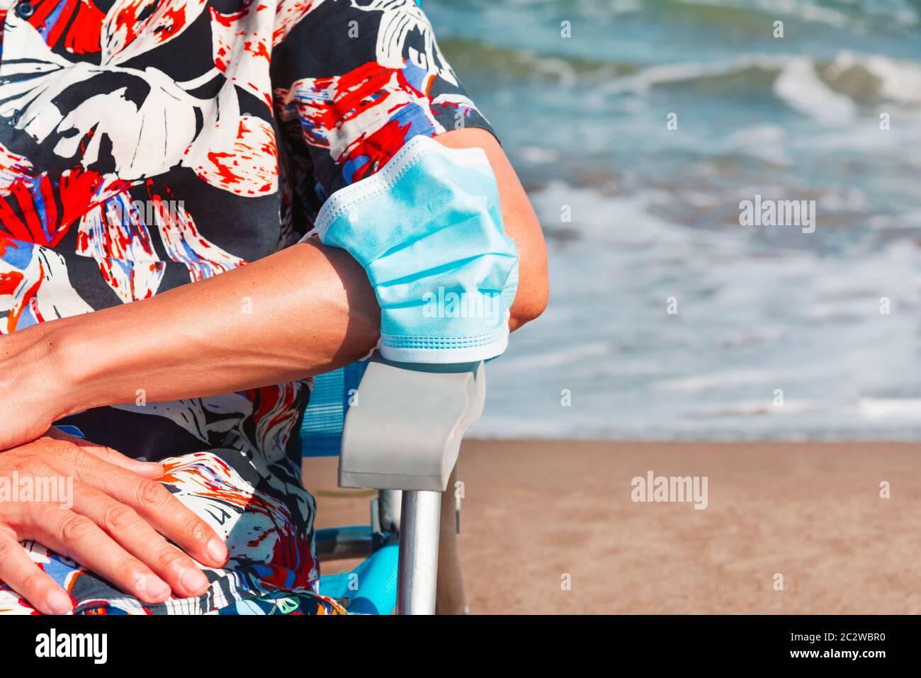 closeup of a caucasian man, sitting in a deck chair on the beach, wearing a blue surgical mask in his arm, as is taking a breather of wearing it, with Stock Photo