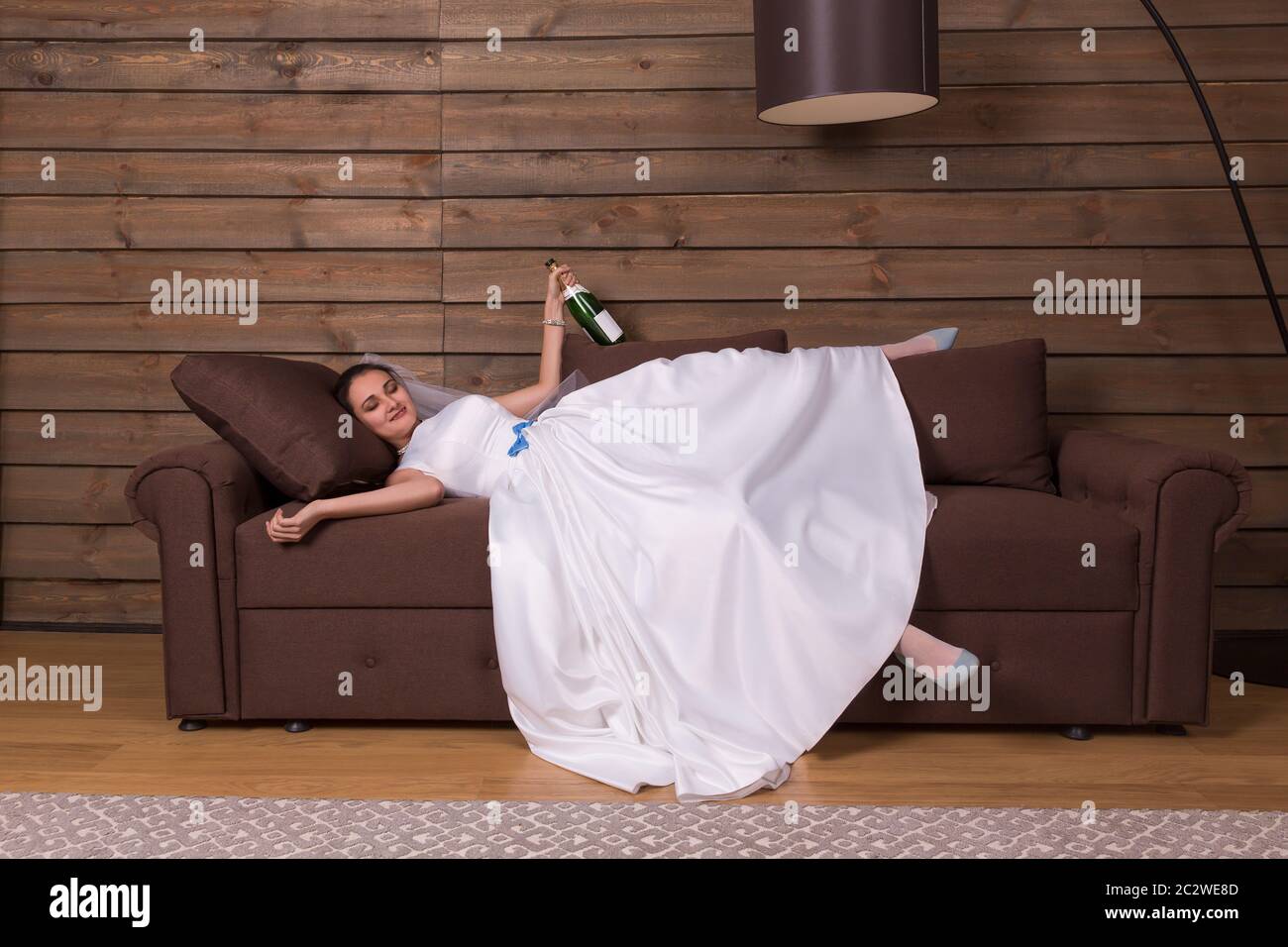 Drunk bride with bottle of alcohol in hand relax on couch after wedding celebration. Wooden interior of the room on background Stock Photo