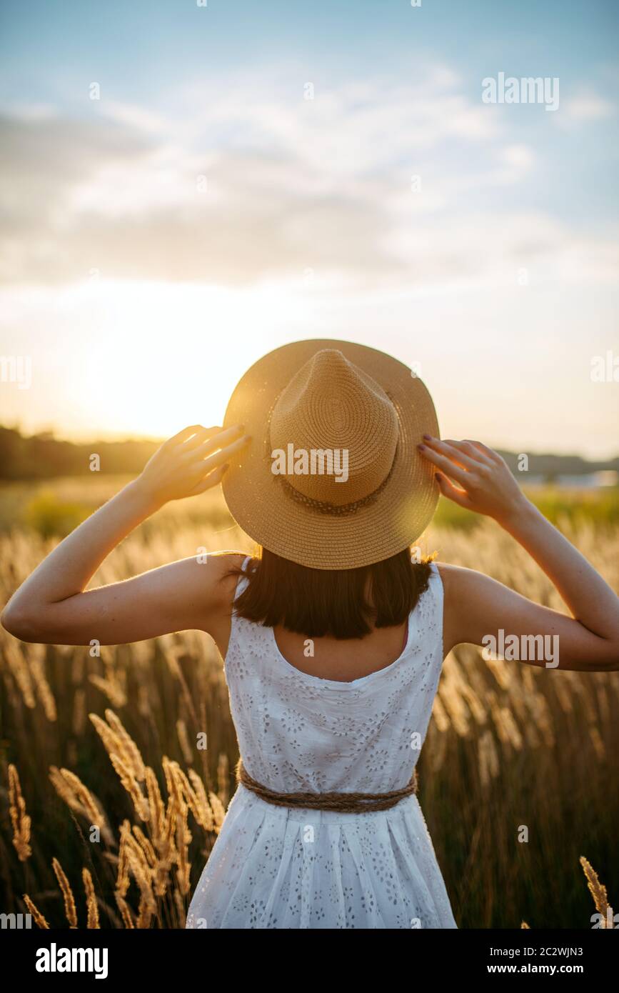 Woman in white dress and straw hat walking in wheat field on sunset ...