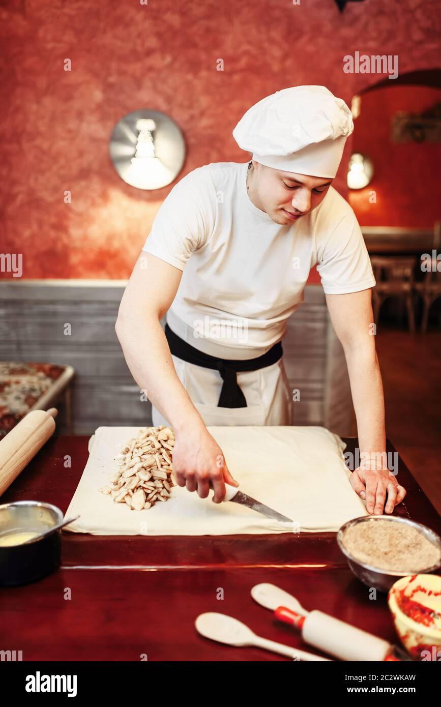 Male chef prepares apple strudel on wooden kitchen table, pastry ingredients on background. Homemade sweet dessert, pie preparation process Stock Photo