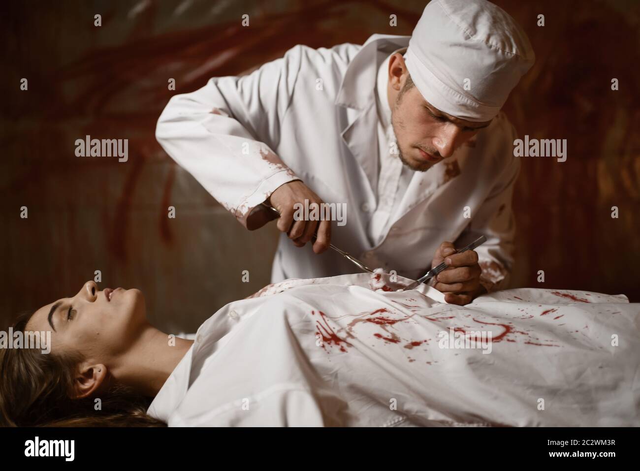 Crazy psychiatrist maniac makes an incision to female patient, experiment in mental hospital basement, bloody walls on background. Victim of mad docto Stock Photo