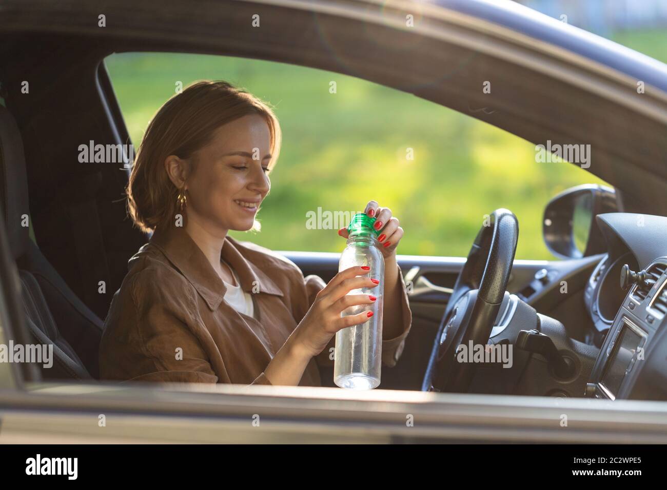 Woman driver drinks water from a refillable bottle in her car, thirsty behind the wheel, stopped to rest. Stock Photo