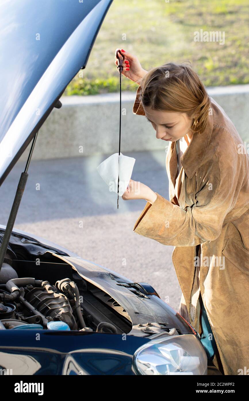 Confident caucasian woman driver checking oil level in car engine dipstick, cityscape on background. Stock Photo