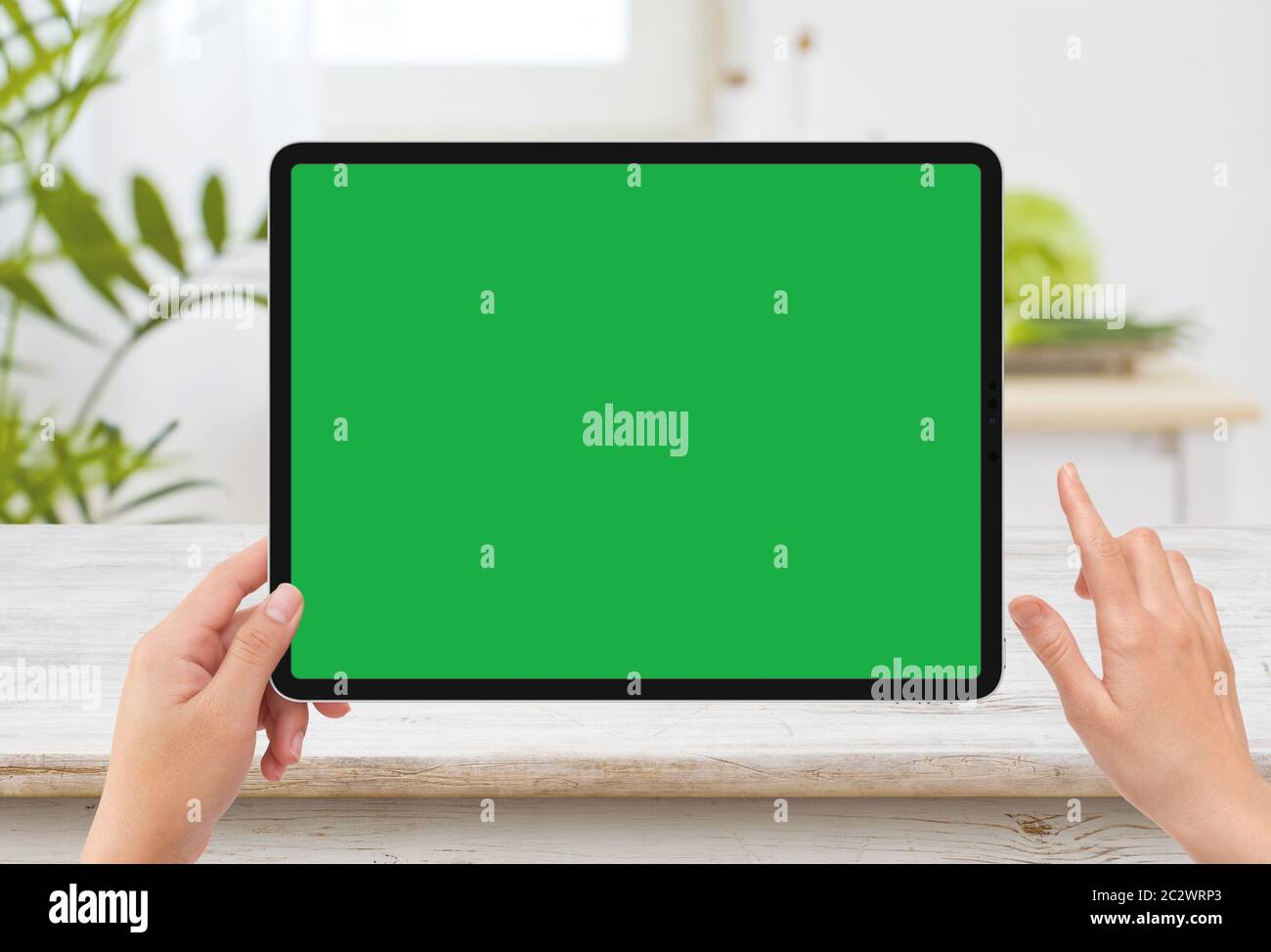 Isolated human left hand holding black tablet media device with white green screen mockup and wooden table in kitchen Stock Photo