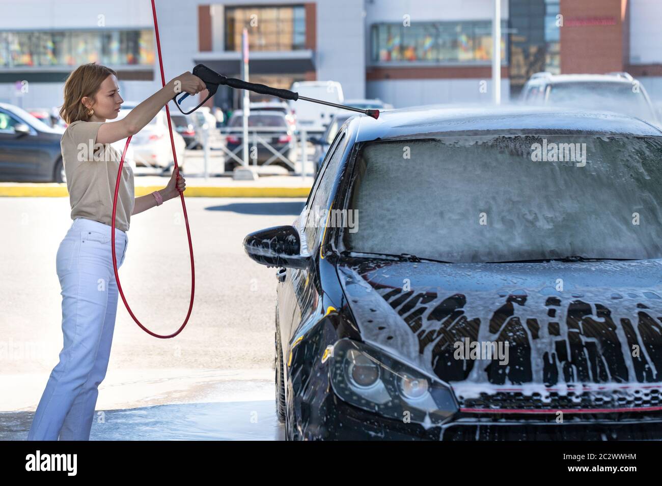 Caucasian young woman driver washing at manual car washing, cleaning with foam, pressured water. Self-service car wash with high-pressure hose Stock Photo