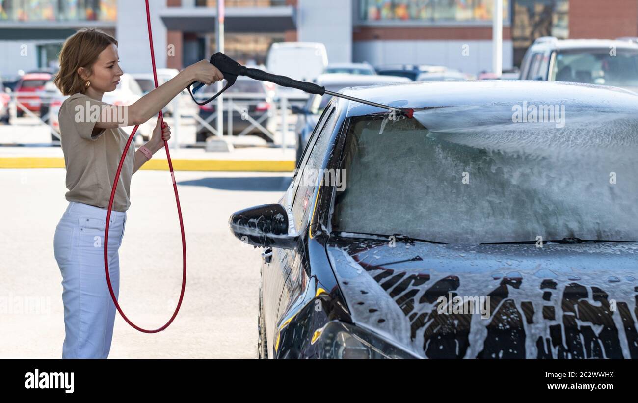 Caucasian young woman driver washing at manual car washing, cleaning with foam, pressured water. Self-service car wash with high-pressure hose Stock Photo