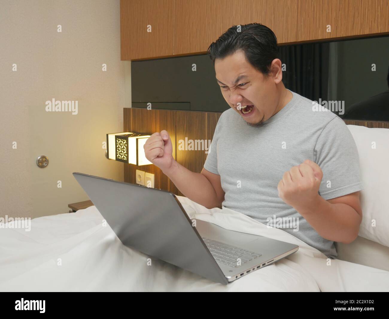 Portrait of young Asian man celebrating victory while looking at laptop on his bed, winning gesture with big smile, entrepreneur working online busine Stock Photo