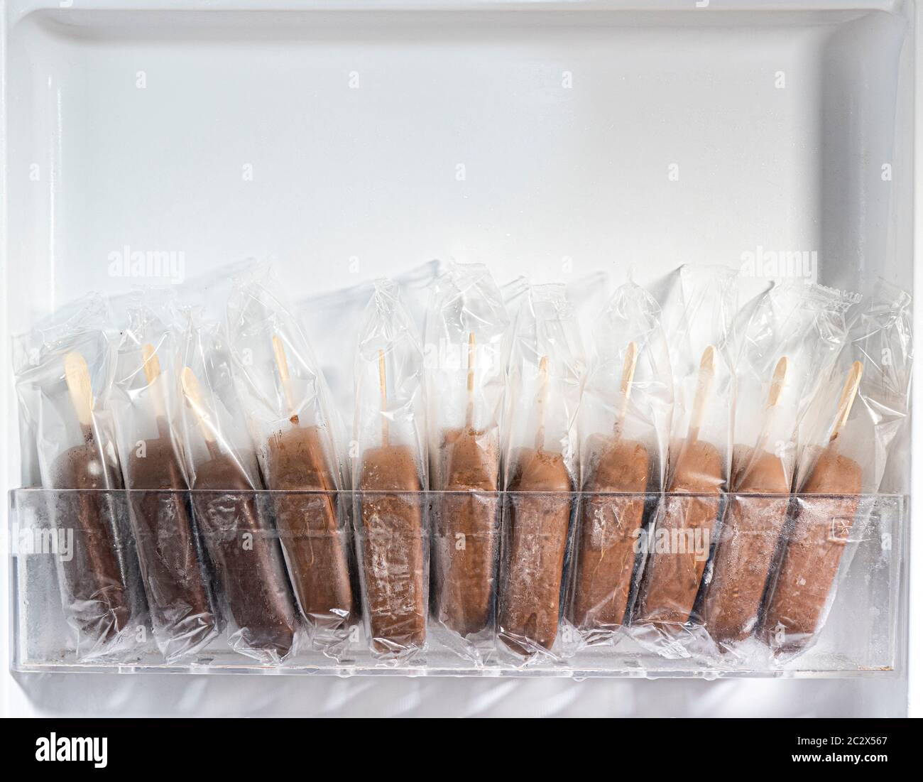 Closeup of 11 unbranded chocolate coated ice cream lollies in transparent wrappers, standing in a row in the open door of a freezer. Stock Photo
